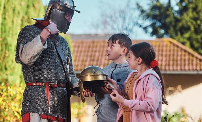 A young girl and boy are standing opposite a man dressed as a Roman gladiator. they are holding a Roman helmet and the gladiator is talking to the chi